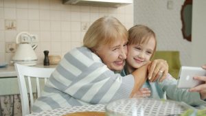 Modern kid girl is taking selfie with loving grandmother using smartphone posing for camera hugging sitting at kitchen table together. Family and gadgets concept.